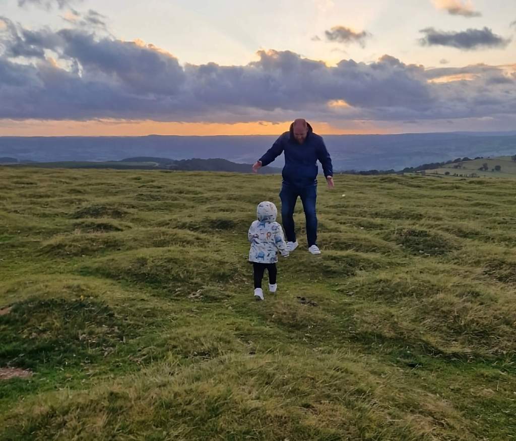 A father and toddler running toward each other across grassy Welsh hills, capturing a joyful family moment