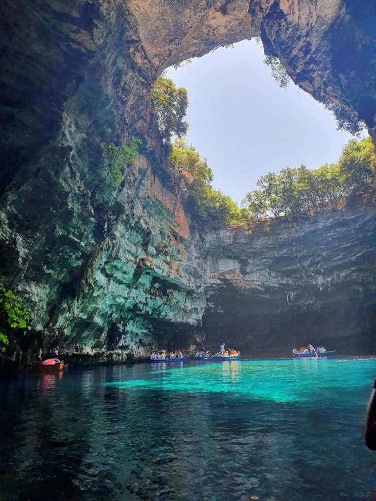 Rowboat gliding through crystal-clear water inside Melissani Cave, sun rays shining through cave roof