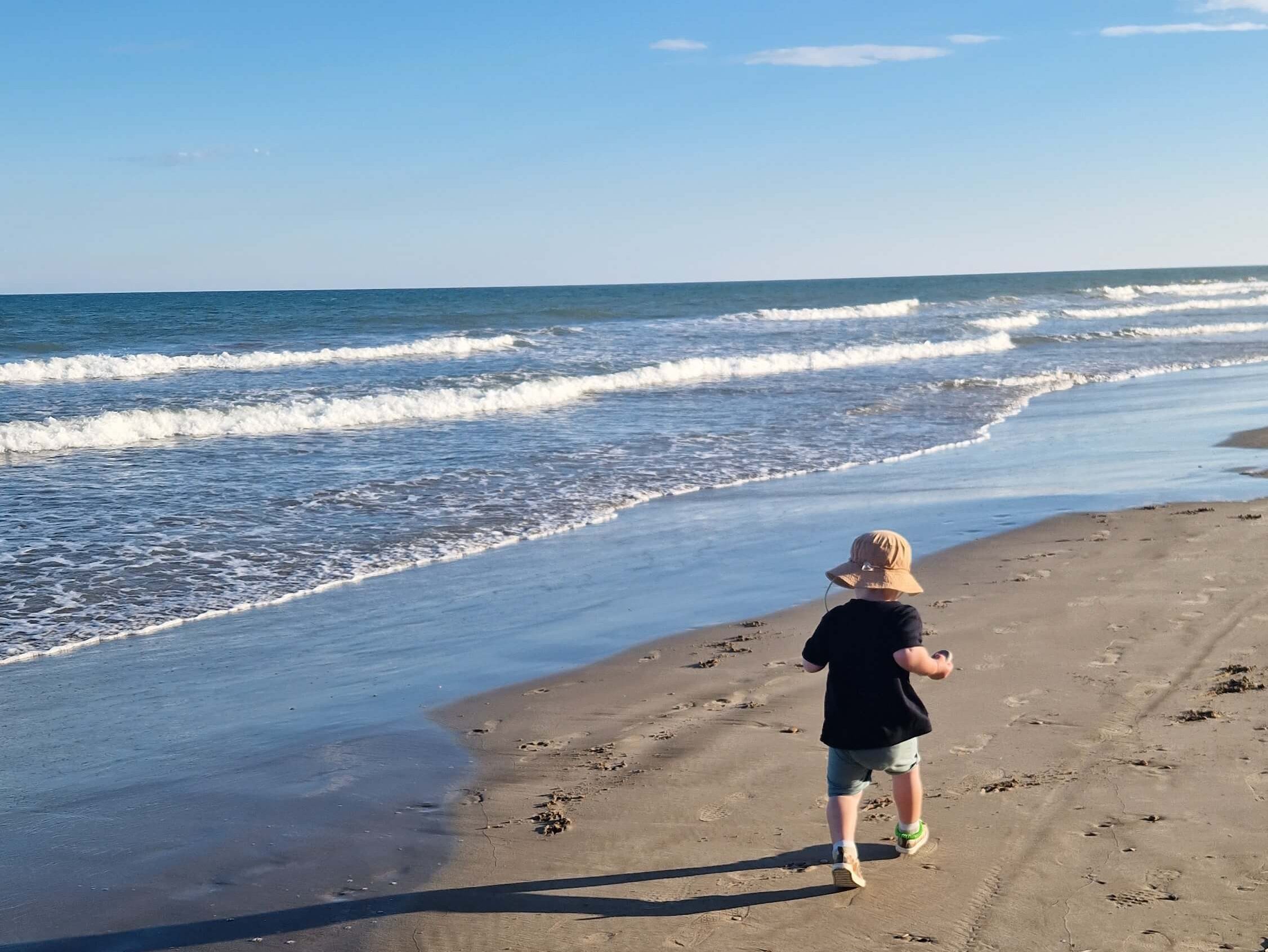 Toddler running along a sunny Spanish beach, back to camera, capturing warm weather vibes.