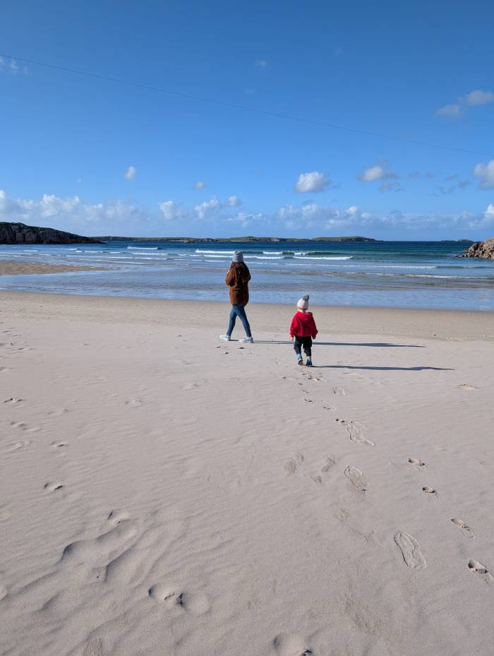 Mum and toddler walking along a white sand beach on the NC500
