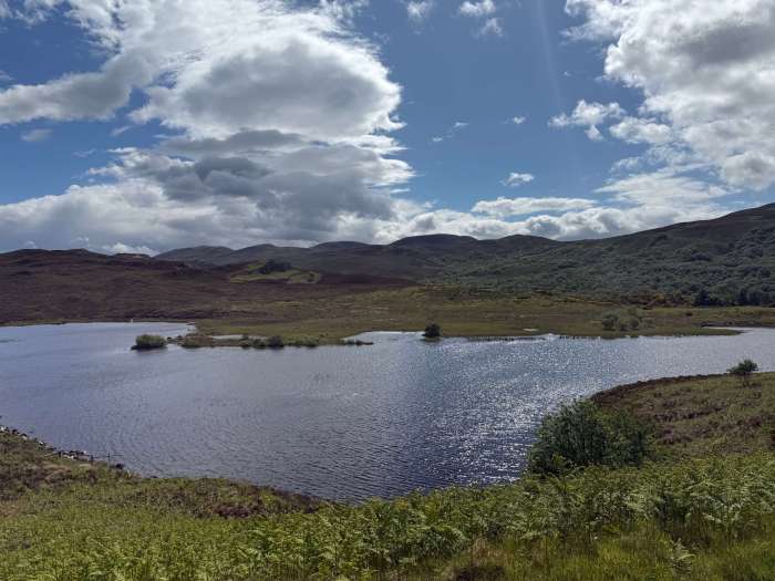 Still loch surrounded by green hills under a sunny Scottish sky