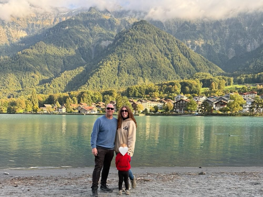 Family of three standing together by a lake in Switzerland with mountains behind